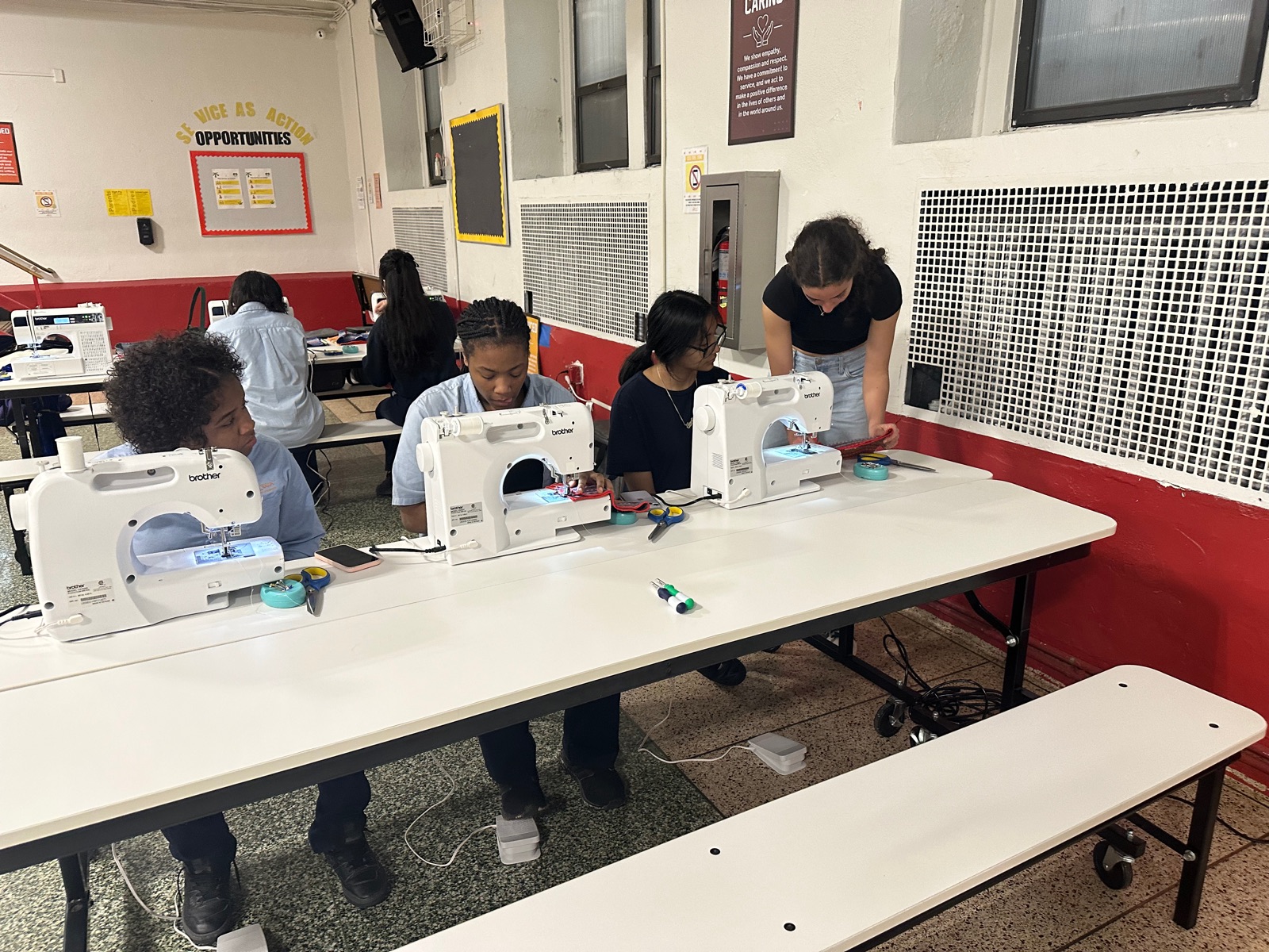A row of students at sewing machines on a long cafeteria table, with Noa helping at the far end