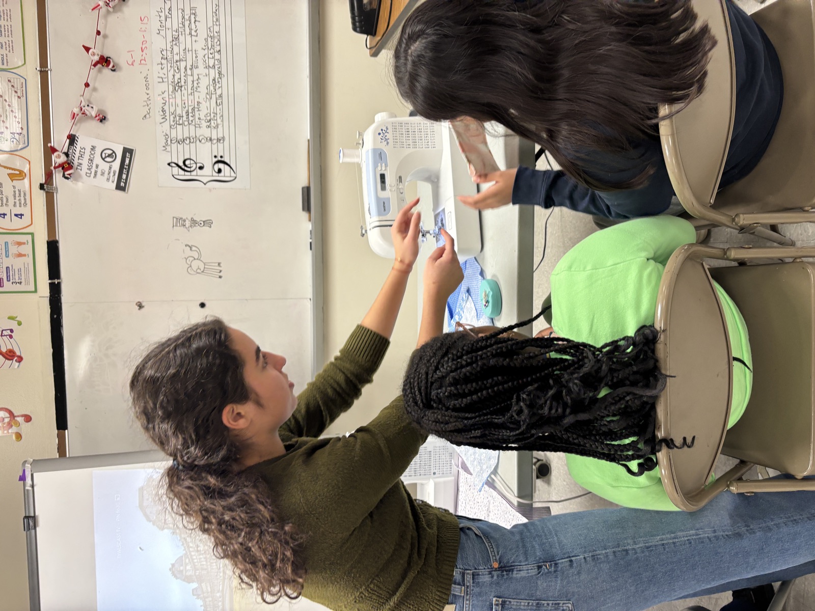 Noa leaning over two students at a sewing machine during a Girls Inc. class